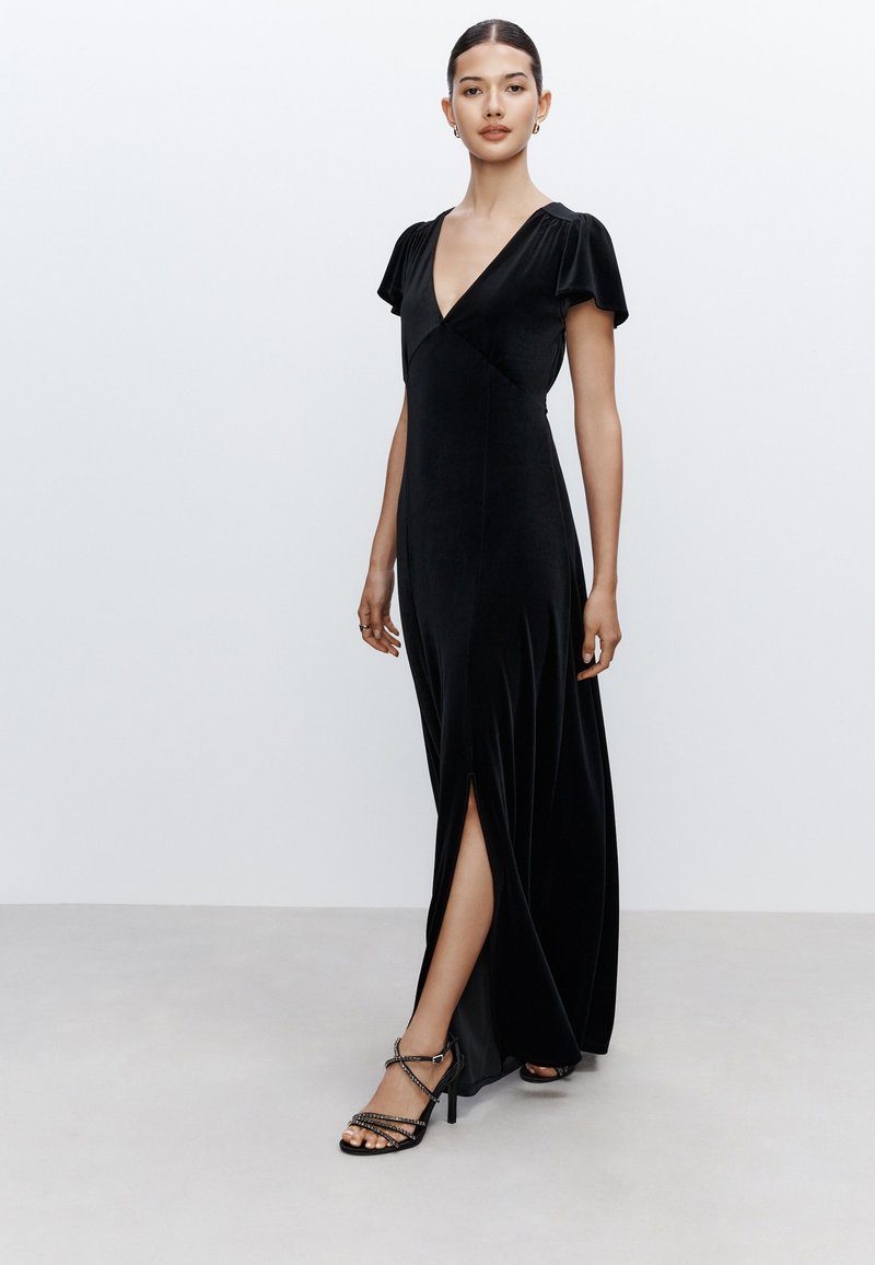 Abito lungo in velluto nero con scollo a V, maniche a svolazzo e spacco laterale. Abbinato a sandali neri con tacco alto e cinturini.