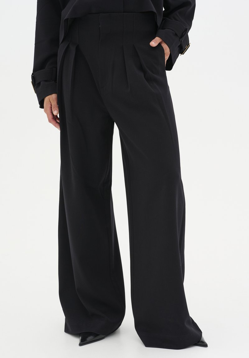 Pantaloni neri a gamba larga con pieghe, caratterizzati da una texture del tessuto liscia e tasche laterali. Progettati con una vita alta e una silhouette sagomata.