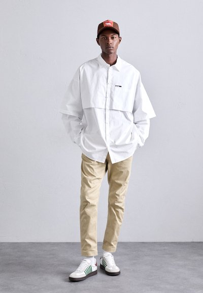 Cămașă albă supradimensionată, cu un design scurtat, pantaloni chino bej și adidași gri cu dungi verzi. O șapcă maro completează look-ul.