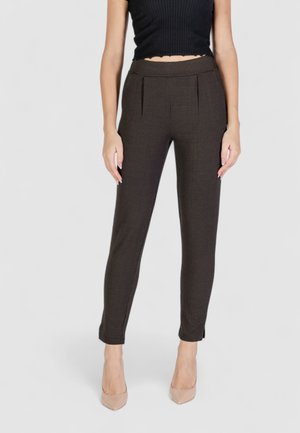 Pantaloni a quadri marroni con una forma aderente, dotati di pieghe frontali, tasche laterali e gambe aderenti. Indossati con tacchi nudi.