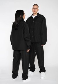 Black oversized jackets made of smooth fabric with snap buttons, paired with matching black pants. Both wear white sneakers.