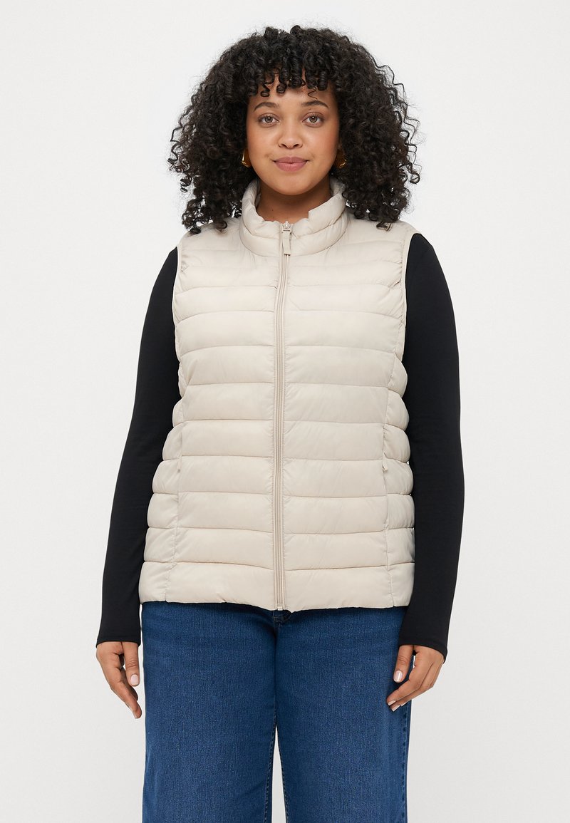 Šviesiai rudos puffer liemenė su aukštu apykakle, horizontaliai suformuotu pamušalu ir priekiniais užtrauktukais. Derinama su ilgomis rankovėmis juodu marškinėliu ir mėlynais džinsais.