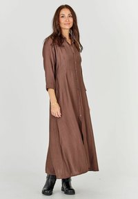 Brown, long-sleeve maxi dress with button-down front and textured fabric. Worn with black ankle boots; dress features a smooth, flowing silhouette.