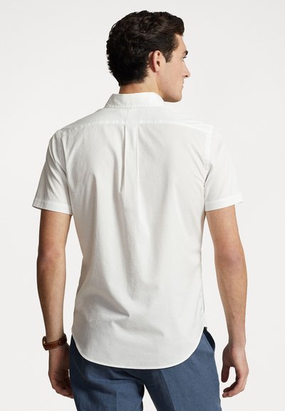 Chemise blanche à manches courtes avec un col, dotée d'un pli au dos et d'un ourlet arrondi ; associée à un pantalon bleu. Texture de tissu lisse.