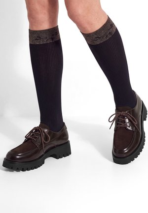 Jambes portant des chaussettes hautes côtelées noires avec des bords scintillants marron et des chaussures épaisses à lacets marron foncé sur fond blanc.