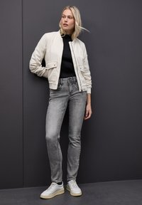 Chaqueta de corduroy beige claro con textura acanalada, dos bolsillos frontales, combinada con jeans gris ajustados y zapatillas blancas. El fondo es gris oscuro.