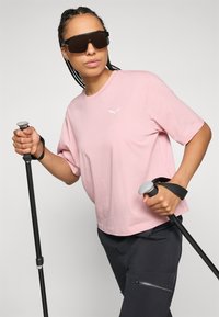 Helle rosa Baumwoll-T-Shirt mit einem weißen Logo, kombiniert mit schwarzen Hosen mit einer Reißverschlusstasche. Das Modell hält Trekkingstöcke.