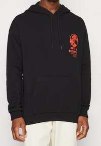 Sudadera de algodón negra con bolsillo frontal, capucha con cordones y texto gráfico rojo en el pecho. Textura suave y corte relajado.