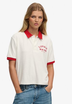 Ung kvinde står med hænderne i lommerne iført en hvid polotrøje med rød krave og manchetter samt blå jeans.