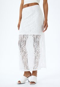 Jupe maxi blanche en dentelle avec des motifs floraux, une texture semi-transparente, une taille ajustée et une silhouette droite. Associée à des sandales à talons blanches.