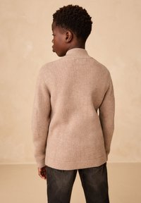 Suéter beige de punto con cuello alto y textura acanalada, combinado con jeans vaqueros oscuros. Vista desde atrás sobre un fondo neutro.