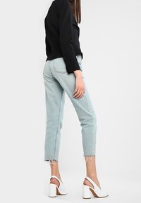 Kvinna som bär ljusblå cropped jeans med fransade kanter, en svart långärmad topp och vita blockklack slingbackskor, stående i profil.