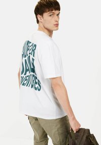 Witte katoen t-shirt met grote teal letters op de achterkant die "UNDER DAILY ADVENTURES" zeggen. Model draagt olijfgroene cargo broek.