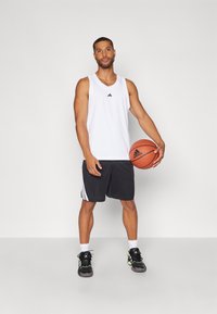 Biely basketbalový top so sieťovinou a čiernym logom, v kombinácii s čiernymi šortkami so bielymi pruhmi. Športovec drží basketbal.