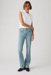 Svetlo modri razširjeni jeans z visokim pasom, v kombinaciji z belo majico brez rokavov. Model pozira z eno roko na boku, stoji v črnih škornjih.