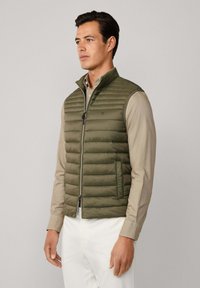 Rompi zaitun, puffer dengan jahitan horizontal, kerah berdiri, dan penutupan ritsleting. Dipakai di atas kemeja lengan panjang beiges muda.