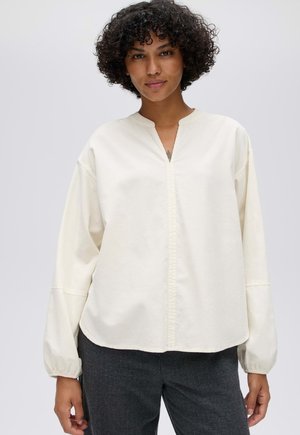 Vrouw met krullend haar die een losse witte blouse met lange mouwen en donkergrijze broek draagt tegen een effen lichte achtergrond.