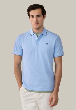 Man draagt een lichtblauw poloshirt met korte mouwen, donkere biezen en logo, gedragen over een groen shirt, gecombineerd met een witte broek, tegen een effen achtergrond.