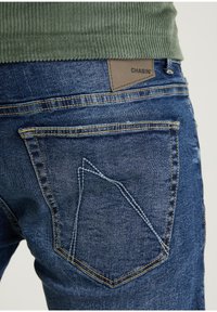 Vaqueros de denim azul con un acabado texturizado, costuras amarillas en contraste y una etiqueta de cuero marrón en la cintura. Combinados con una parte superior verde de canalé.