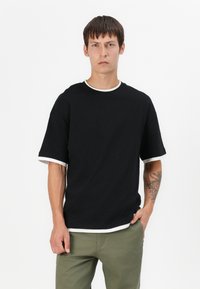 Black short-sleeve T-shirt with a round neckline and white trim at the collar and sleeves, made from a soft fabric. Worn with green pants.