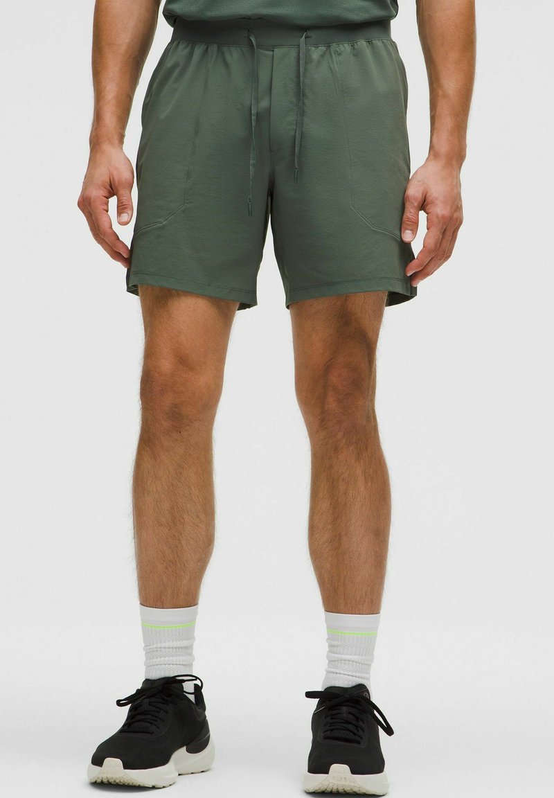 Shorts sportivi verdi con vita elastica, chiusura con lacci, e due tasche laterali, abbinati a sneakers nere e calze bianche.