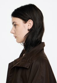 Giacca marrone con colletto ampio, chiusura con zip e texture liscia. Il modello ha i capelli lunghi e neri e indossa un piccolo orecchino a cerchio nero.