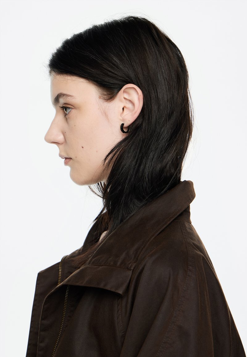 Giacca marrone con colletto ampio, chiusura con zip e texture liscia. Il modello ha i capelli lunghi e neri e indossa un piccolo orecchino a cerchio nero.