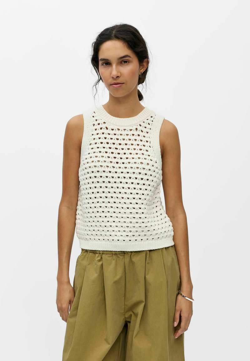 Femme aux cheveux foncés en queue de cheval basse, portant un haut blanc en crochet sans manches et un pantalon ample vert olive sur un fond uni.