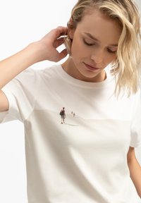 Witte katoenen t-shirt met een subtiele landschapsafbeelding van twee figuren die op het zand lopen, ontworpen met een ronde hals en korte mouwen.