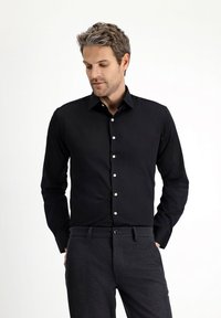 Czarna koszula na długi rękaw z zapięciem na guziki z przodu, spiczastym kołnierzykiem i białymi guzikami, zestawiona z ciemnoszarymi spodniami. Gładka, jednolita tkanina.