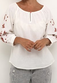 Femme portant un chemisier blanc avec des broderies florales brunes sur les manches et un bouton à l'avant, associé à un jean gris clair déchiré.