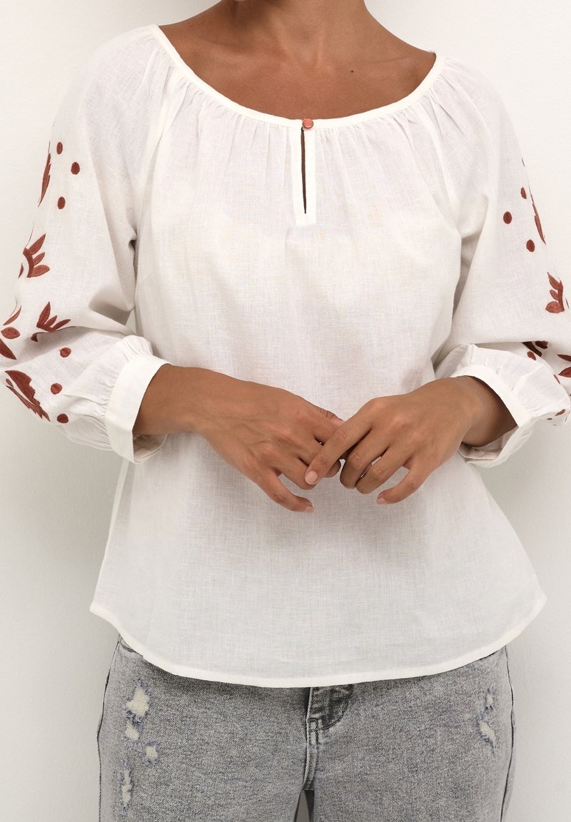 Femme portant un chemisier blanc avec des broderies florales brunes sur les manches et un bouton à l'avant, associé à un jean gris clair déchiré.