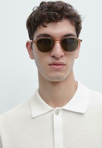 Occhiali da sole rotondi con lenti color marrone e montatura sottile di colore chiaro. Il modello indossa una polo bianca con trama e patta con bottoni.