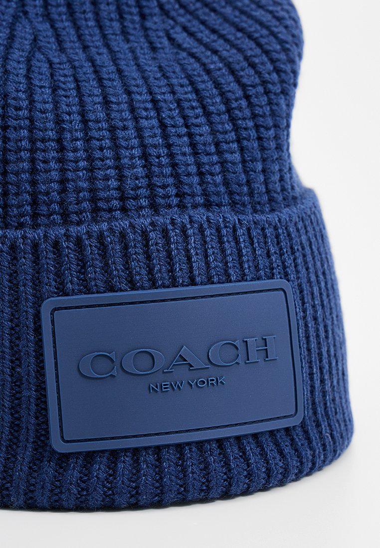 Navy blue knitted beanie featuring vertical ribbing, a textured finish, and a raised, rectangular rubber patch with "COACH NEW YORK" logo.