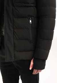 Giacca a vento nera in tessuto trapuntato, con tasca con zip e polsini a costine. Presenta una zip resistente e un design elegante e minimalista.