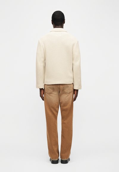 Άτομο που φοράει κρεμ πλεκτό πουλόβερ με ρίγες, καφέ παντελόνι ίσιας γραμμής και μαύρα παπούτσια, στέκεται με την πλάτη προς τα έξω σε απλό λευκό φόντο.