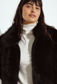 Melnas mākslīgās kažokādas apvalks ar spīdīgu tekstūru, apvienots ar krēmkrāsas augstu kaklu, kuram ir ribotu detaļu akcenti. Tuvplāns.