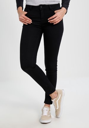 Osoba nosząca czarne obcisłe dżinsy i beżowe trampki, stojąca z jedną nogą skrzyżowaną nad drugą i rękami spoczywającymi na biodrach.