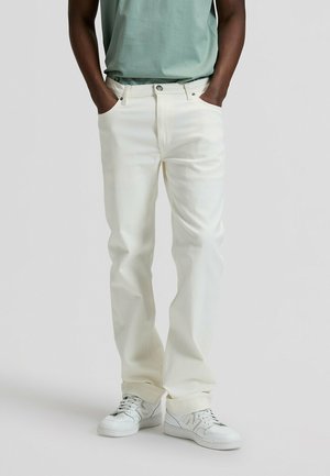 Person iført cremefarvede jeans med lige ben, med hænderne i lommerne og hvide sneakers mod en ensfarvet baggrund.