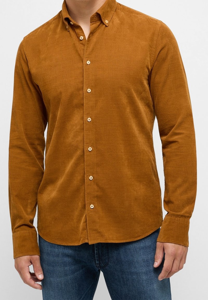 Camicia button-up in cotone corduroy marrone con colletto, maniche lunghe e chiusura frontale a bottoni, abbinata a jeans in denim blu.
