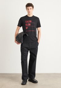 Czarna bawełniana koszulka z czerwonym napisem "CALVIN KLEIN NY." Połączona z czarnymi spodniami i trzymana czarna torba na akcesoria. Prosty design.