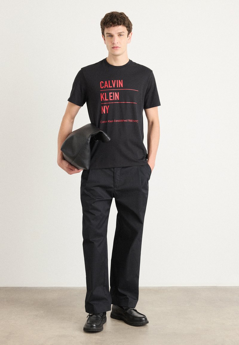 Camiseta de algodón negra con texto rojo que dice "CALVIN KLEIN NY." Combinada con pantalones negros y sosteniendo un bolso de accesorios negro. Diseño sencillo.