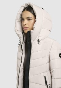 Light pink puffer jacket with a large hood, featuring a black zipper and accents. Quilted texture with branding on the zipper and sleeve.