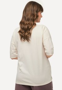 Cremefarvet kortærmet T-shirt med afslappet pasform, rundet kant og sideslids, lavet af blødt, let stof. Vist som bæret fra ryggen.