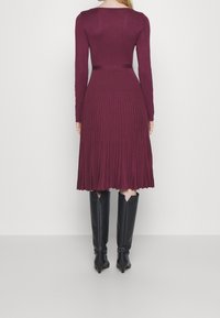 Robe bordeaux à manches longues et jupe plissée, dotée d'un haut ajusté et d'une texture lisse. Portée avec des bottes noires montantes jusqu'au genou.