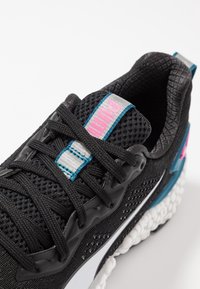 Gros plan sur une chaussure de sport Puma noire avec un dessus en mesh, des lacets noirs, un logo rose sur la languette, une semelle blanche et des accents teal.
