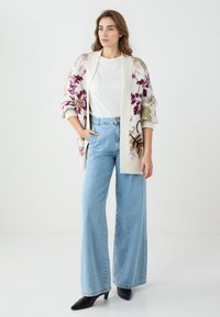 Übergroßer cremefarbener Cardigan mit Blumendessin in Lila und Grün, weißes T-Shirt, weit geschnittene blaue Jeans und schwarze spitze Pumps.