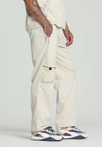 Pantaloni cargo beige chiaro con dettagli sulle tasche laterali, vestibilità ampia e una texture sottile. Abbinati a sneakers colorate con molteplici colori d'accento.
