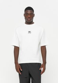 Hvid T-shirt med korte ærmer lavet af bomuld, med et lille sort myre grafisk design og mærkenavnet "Oakley" i midten. Glat tekstur.