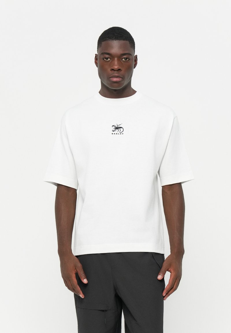 Hvid T-shirt med korte ærmer lavet af bomuld, med et lille sort myre grafisk design og mærkenavnet "Oakley" i midten. Glat tekstur.